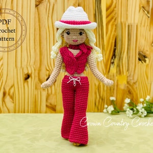 Könnte beinhalten: Gehäkelte Puppe mit weißem Hut mit rosa Band, rosa Weste und passender Hose. Die Puppe hat blondes Haar und steht vor einem Holz Hintergrund. Der Text "PDF Crochet Pattern" befindet sich in einem Kreis.