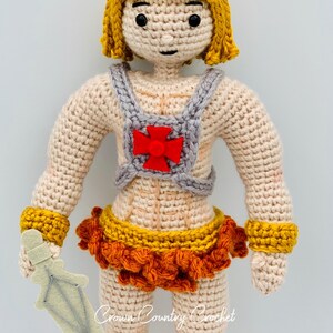 May include: Crocheted doll with blonde hair, a red cross on its chest, and a sword. The doll is wearing a grey vest, a yellow and orange skirt, and yellow bracelets. The doll is holding a white sword.