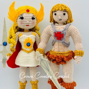 May include: Crocheted figures of He-Man and She-Ra, both with yellow hair and wearing their iconic costumes. He-Man is holding a sword and She-Ra is holding a sword and shield. The figures are standing on a white background.