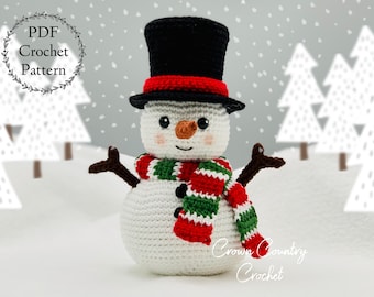 May include: A crocheted snowman wearing a black top hat with a red band, a green and red striped scarf, and a white body. The snowman has black eyes, a brown nose, and a black button mouth. The background is a snowy scene with white trees.