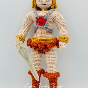 May include: Crocheted doll with blonde hair, wearing a red and gray chest piece, orange skirt, and orange boots. The doll is holding a white sword.