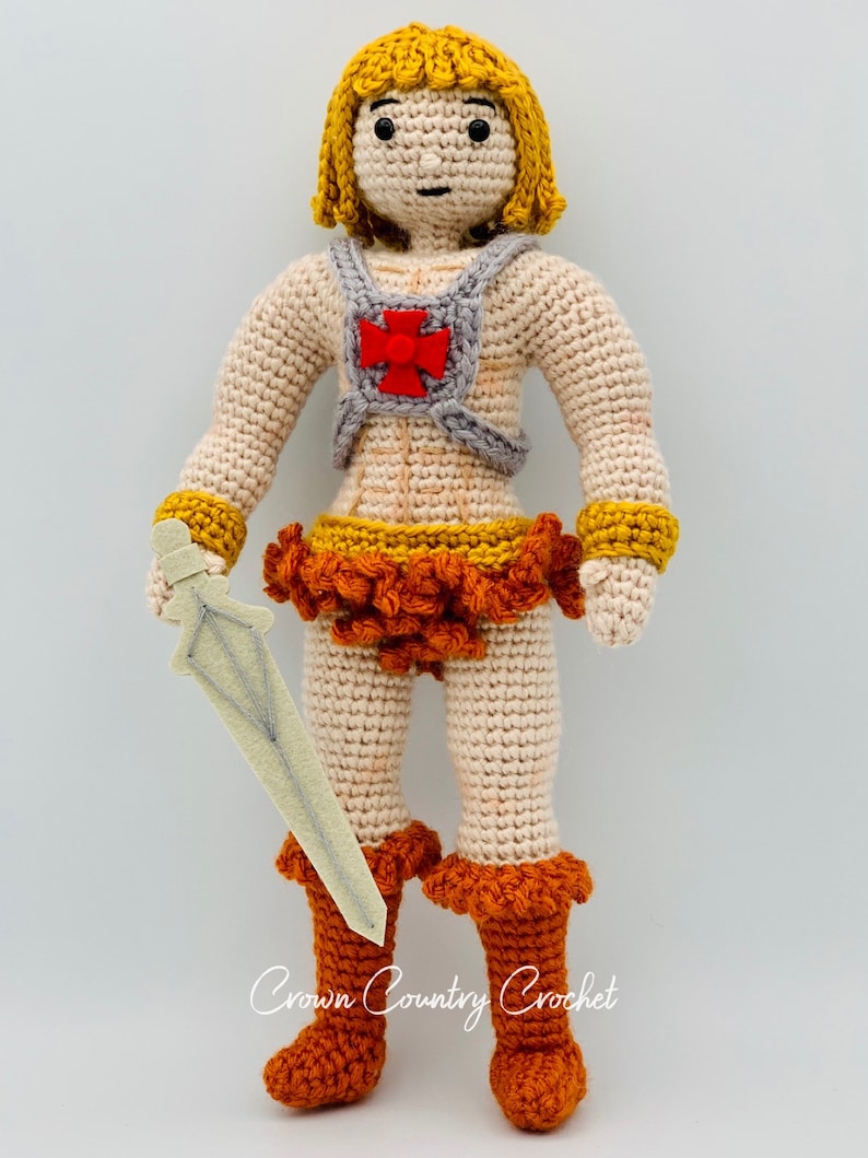 May include: Crocheted doll with blonde hair, wearing a red and gray chest piece, orange skirt, and orange boots. The doll is holding a white sword.