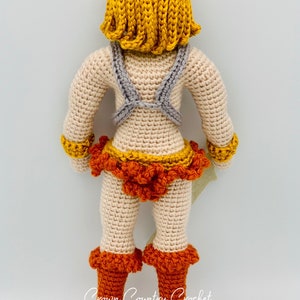 May include: Crocheted doll with a yellow wig, gray chest armor, orange and yellow skirt, and orange boots. The doll is standing on a white background. The text "Crown Country Crochet" is visible at the bottom of the image.
