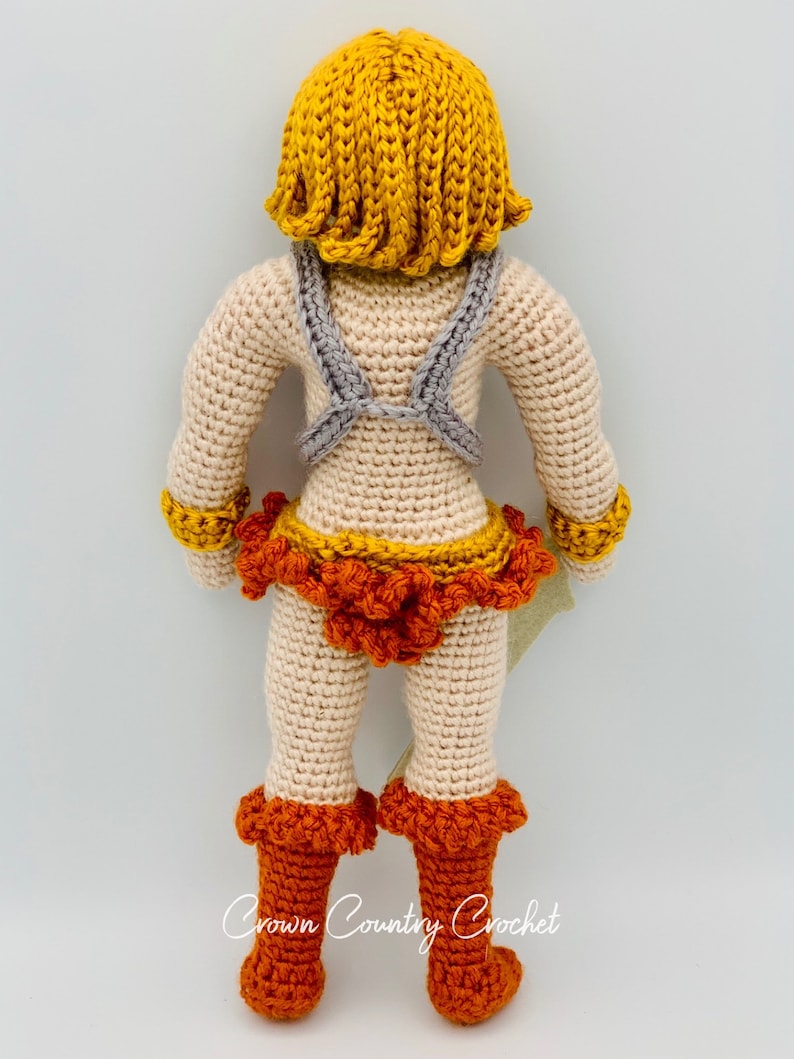 May include: Crocheted doll with a yellow wig, gray chest armor, orange and yellow skirt, and orange boots. The doll is standing on a white background. The text "Crown Country Crochet" is visible at the bottom of the image.