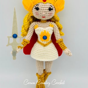 May include: A crocheted doll of a female character with blonde hair, wearing a white dress, yellow boots, a yellow crown, and holding a silver sword. The doll is standing on a white background.