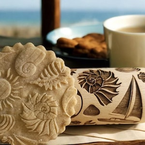 Könnte beinhalten: Ein Holz-Nudelholz mit einem Muschel- und Segelboot-Design. Das Nudelholz wird verwendet, um dekorative Muster auf Teig zu erstellen. Ein Keks mit einem ähnlichen Design befindet sich im Vordergrund.