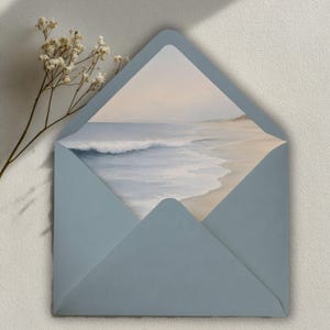 Könnte beinhalten: Ein staubblauer Umschlag mit dreieckiger Klappe, der eine Karte mit einem sanften Aquarellgemälde von einem Sandstrand, Meereswellen und einem blassen Himmel enthält. Ein zarter Zweig mit getrockneten weißen Blumen liegt neben dem Umschlag.