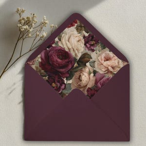 Può includere: Una busta color prugna con fodera interna floreale. La fodera presenta un motivo di rose nei toni del bordeaux, crema e rosa. La busta è aperta, rivelando il disegno floreale. Un rametto di fiori bianchi è nell'angolo in alto a sinistra.