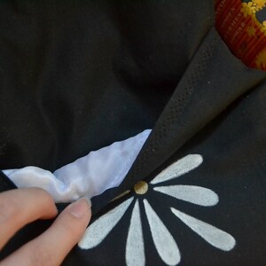 Pode incluir: Tecido preto com um desenho floral branco. O tecido est&aacute; dobrado, revelando um interior branco. Um bot&atilde;o dourado est&aacute; no centro do desenho floral. O item parece ser uma bolsa ou uma bolsa.