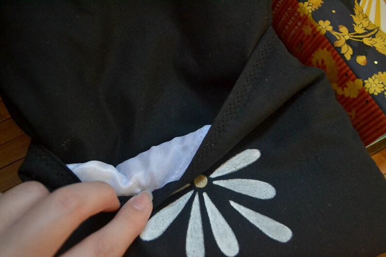 Pode incluir: Tecido preto com um desenho floral branco. O tecido est&aacute; dobrado, revelando um interior branco. Um bot&atilde;o dourado est&aacute; no centro do desenho floral. O item parece ser uma bolsa ou uma bolsa.