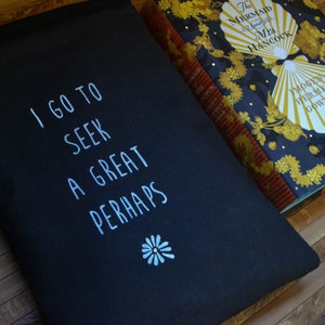 Pode incluir: Bolsa preta com o texto branco "I GO TO SEEK A GREAT PERHAPS". Uma pequena flor branca est&aacute; abaixo do texto. Um livro com capa azul marinho e detalhes dourados est&aacute; ao lado da bolsa.