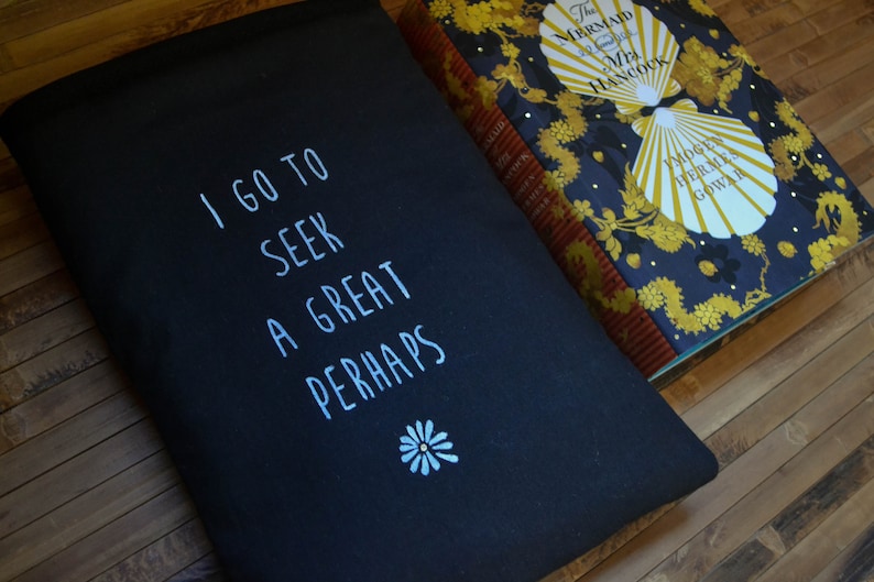 Pode incluir: Bolsa preta com o texto branco "I GO TO SEEK A GREAT PERHAPS". Uma pequena flor branca est&aacute; abaixo do texto. Um livro com capa azul marinho e detalhes dourados est&aacute; ao lado da bolsa.