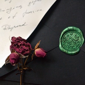 Puede incluir: Primer plano de un sobre negro con un sello de cera verde con un diseño de montaña, luna y estrellas. También se ven una rosa burdeos seca y una nota manuscrita. El sello está en relieve.