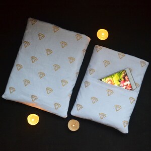 May include: Two white pillows with a gold diamond pattern. One pillow has a pocket with a smartphone inside. The pillows are on a black surface, with three lit candles nearby. The pillows are a decorative item.