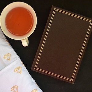 May include: A still-life image featuring a white teacup filled with a reddish-orange liquid, a closed brown book with gold trim, and a white cloth with a gold diamond pattern. The items are arranged on a dark surface.