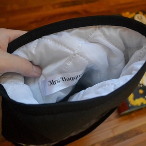 Pode incluir: Bolsa preta com interior acolchoado branco. O interior tem uma etiqueta que diz "Mrs. Baggins". A bolsa &eacute; mantida aberta, revelando o interior. Um livro com um design amarelo e preto &eacute; vis&iacute;vel no fundo.