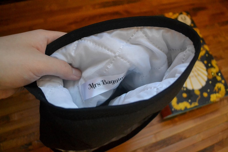 Pode incluir: Bolsa preta com interior acolchoado branco. O interior tem uma etiqueta que diz "Mrs. Baggins". A bolsa &eacute; mantida aberta, revelando o interior. Um livro com um design amarelo e preto &eacute; vis&iacute;vel no fundo.