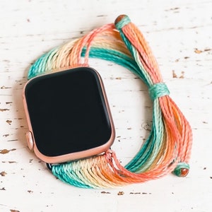 May include: A smartwatch with a black face and a rose gold band. The band is made of a colourful, woven string with shades of pink, orange, yellow, green, and blue.