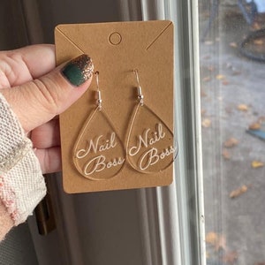 Könnte beinhalten: Ein Paar durchsichtige Acryl-Tropfenohrringe mit dem Schriftzug "Nail Boss" in Weiß.