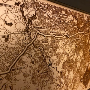 May include: A laser-engraved wooden map of the Boston area, showing the towns of Weston, Waltham, Watertown, Auburndale, Newton, Waban, Wellesley, and Needham. The map is framed in a black frame.