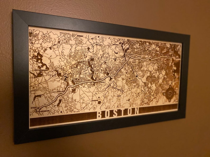 May include: A framed wood map of Boston, Massachusetts. The map is laser engraved with the city's streets and landmarks. The frame is black and the map is a light brown color. The word "BOSTON" is engraved at the bottom of the map.