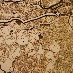 May include: A close-up of a wooden map of Boston, Massachusetts, with the Charles River, Newton, Brookline, and Heartbreak Hill labeled. The map is engraved with brown lines representing streets and roads.