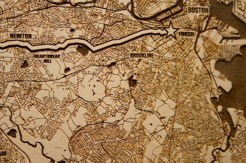 May include: A close-up of a wooden map of Boston, Massachusetts, with the Charles River, Newton, Brookline, and Heartbreak Hill labeled. The map is engraved with brown lines representing streets and roads.