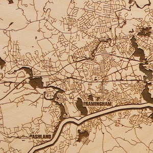May include: A laser-cut wooden map of the Framingham, Massachusetts area. The map shows the towns of Framingham, Ashland, Southborough, and Natick. The map is made of light brown wood and has a detailed design.