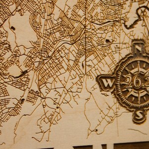 May include: A close-up of a wooden map with a compass design. The compass features the text "26.2" and the directions "N", "S", "E", and "W". The map shows the area around Dedham, Massachusetts.