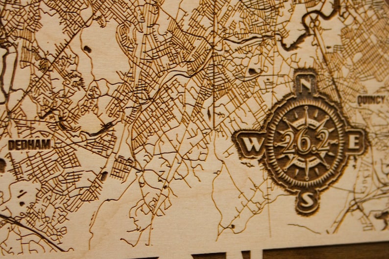 May include: A close-up of a wooden map with a compass design. The compass features the text "26.2" and the directions "N", "S", "E", and "W". The map shows the area around Dedham, Massachusetts.