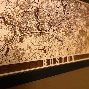 May include: A wooden map of Boston, Massachusetts, with the city name engraved at the bottom. The map is framed in black and has a light brown background.