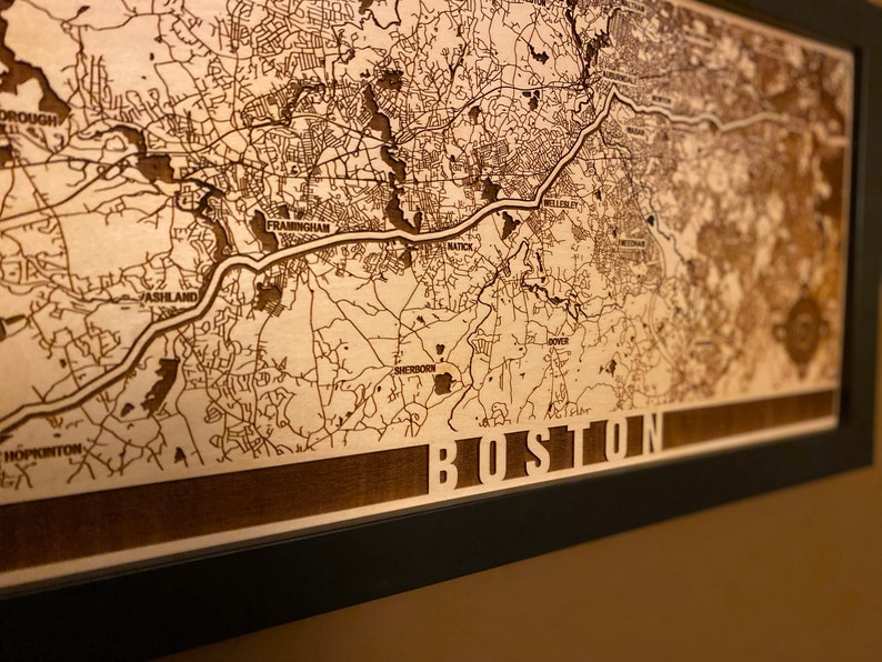 May include: A wooden map of Boston, Massachusetts, with the city name engraved at the bottom. The map is framed in black and has a light brown background.