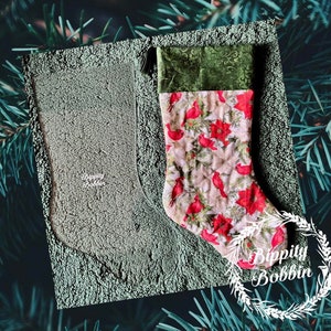 May include: A red and white Christmas stocking with a green velvet cuff and a floral pattern. The stocking is on a green fuzzy surface with the text "Blippity Bobbin" in white.