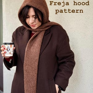 May include: A brown hooded scarf and matching scarf are worn with a brown coat. The text "Freja hood pattern" is displayed in the upper right corner. The person is holding a coffee cup.