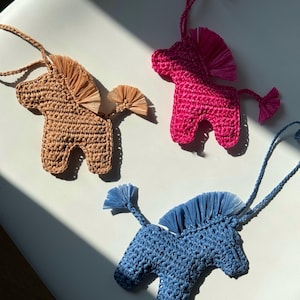 May include: Three crochet horse ornaments in tan, hot pink, and blue. Each horse has a braided cord for hanging, a fringed mane, and a fringed tail. The ornaments are displayed on a white surface with sunlight.
