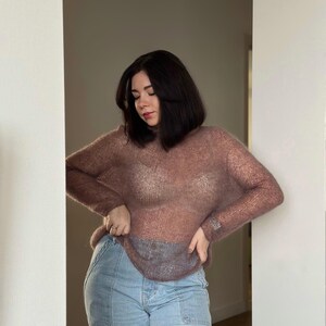 May include: A woman wearing a sheer, mauve sweater over a white vest top and light blue jeans.