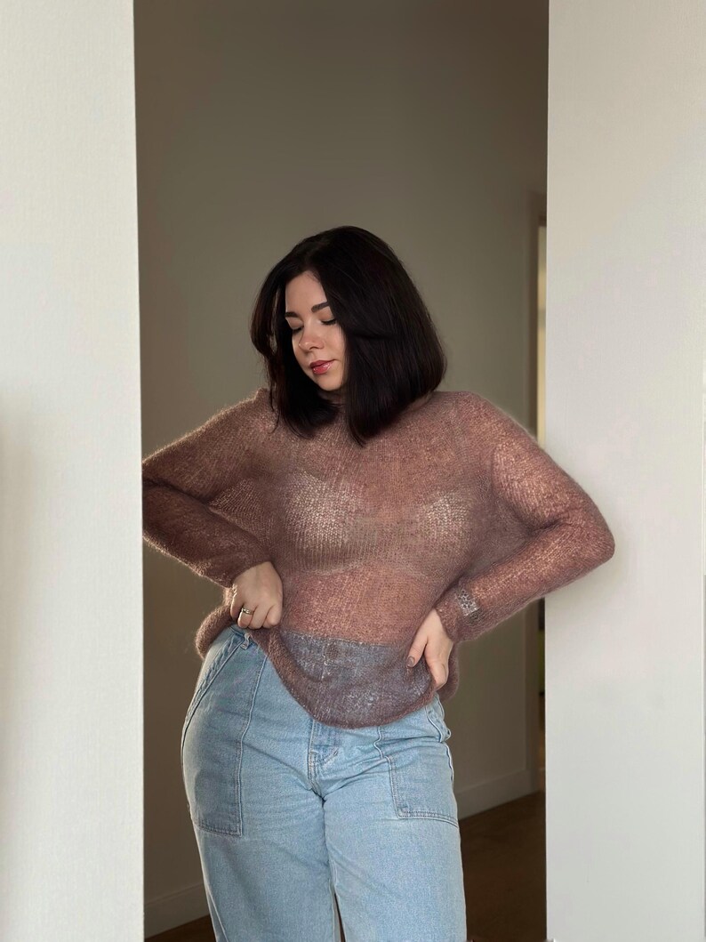 May include: A woman wearing a sheer, mauve sweater over a white vest top and light blue jeans.