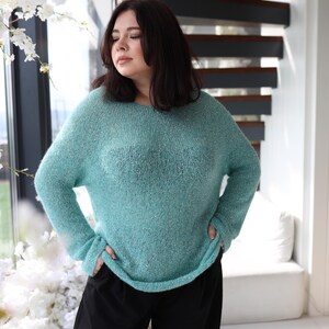 May include: A teal-coloured knitted sweater with a relaxed fit and long sleeves. The sweater has a textured finish and is worn with black trousers. The background features a spiral staircase and white floral displays.