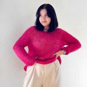 May include: A woman wearing a bright pink fluffy jumper and white trousers. She is standing in front of a white wall.