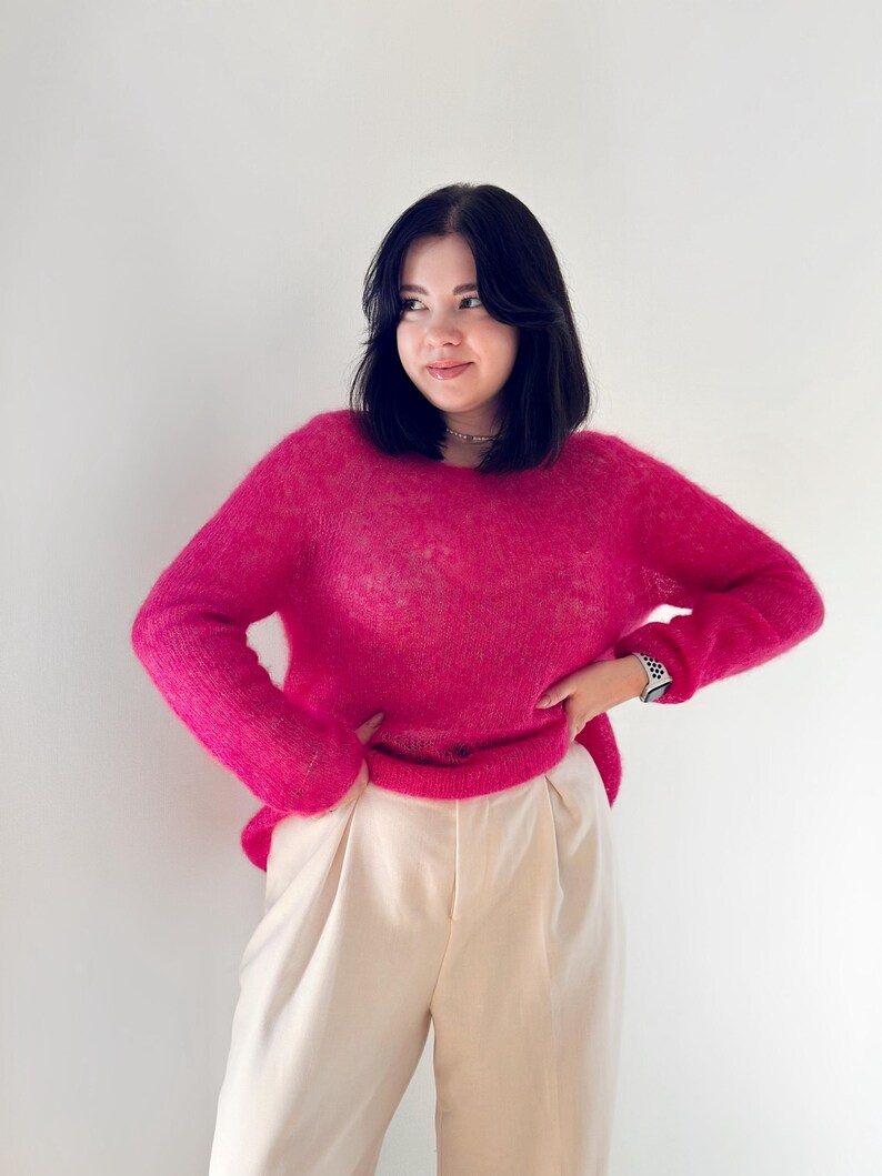 May include: A woman wearing a bright pink fluffy jumper and white trousers. She is standing in front of a white wall.
