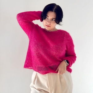 May include: A woman wearing a bright pink fuzzy sweater and beige trousers. She is standing in front of a white background.