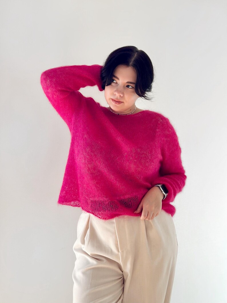 May include: A woman wearing a bright pink fuzzy sweater and beige trousers. She is standing in front of a white background.