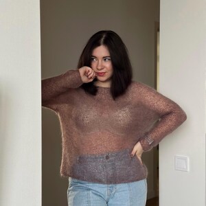 May include: A woman wearing a sheer, mauve-coloured sweater over a white bra and light blue jeans. She is standing in a doorway with her hand on her hip.