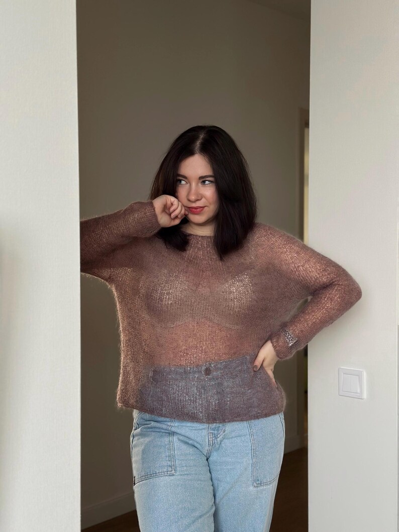 May include: A woman wearing a sheer, mauve-coloured sweater over a white bra and light blue jeans. She is standing in a doorway with her hand on her hip.