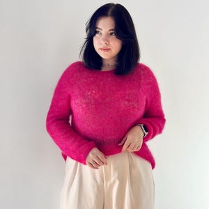 May include: A woman wearing a bright pink fluffy sweater and cream-coloured trousers. She is standing in front of a white wall.