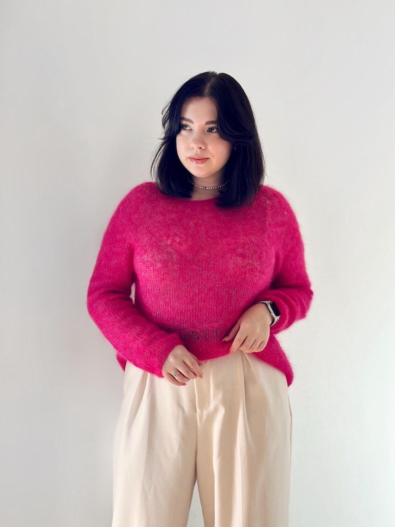 May include: A woman wearing a bright pink fluffy sweater and cream-coloured trousers. She is standing in front of a white wall.