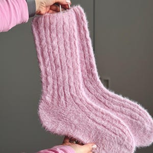 Könnte beinhalten: Ein Paar weiche, hellrosa flauschige Socken mit Zopfmuster. Die Socken werden von einer weißen Schnur gehalten. Die Socken bestehen aus einem flauschigen Material und wirken warm und gemütlich.