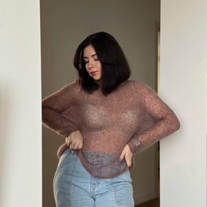 May include: A woman wearing a sheer, mauve sweater over a white tank top and light blue jeans.
