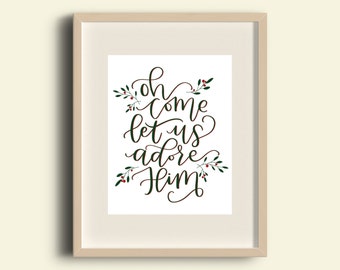 May include: A framed print with a white background and green calligraphy that reads "Oh come let us adore Him". The calligraphy is surrounded by green leaves with red berries.