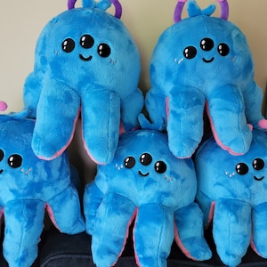 Puede incluir: Cinco peluches de pulpo azules con detalles rosas y tres ojos negros cada uno. Los peluches de pulpo están dispuestos en forma de pirámide.